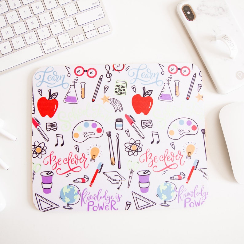 May include: A white mouse pad with a colorful pattern of school supplies, including apples, pencils, paintbrushes, and musical notes. The text "Learn", "Be Clever", and "Knowledge is Power" is written on the mouse pad.
