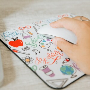 May include: White mouse pad with a black border featuring a colorful doodle design of school supplies, including an apple, paintbrushes, a globe, and the words "Learn", "School", and "Be Clever".