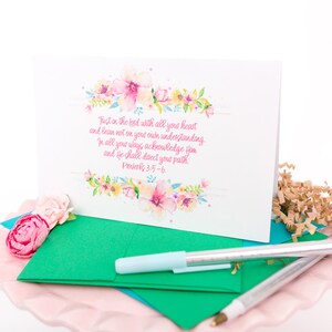 May include: A white greeting card with floral watercolor accents and the text "Trust in the lord with all your heart... Proverbs 3:5-6." A green envelope, pink flower, and pens are also visible.