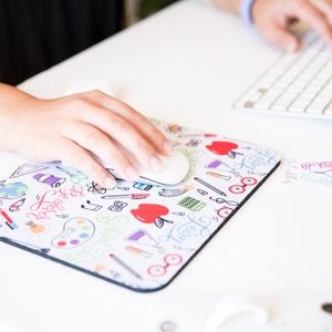 May include: A white computer mouse resting on a colorful mousepad with a school-themed design. The mousepad features a variety of colorful doodles, including apples, pencils, and a calculator. The text "Learn" is written in a whimsical font on the mousepad.