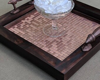 May include: A decorative wooden serving tray with a copper-colored mosaic tile center and dark brown frame. It features bronze handles and a clear glass dish filled with white flower petals.