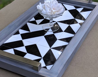 May include: A decorative serving tray with a geometric black and white tile design. The tray has a gray wooden frame and silver handles. A clear glass dish with white flowers sits on the tray.