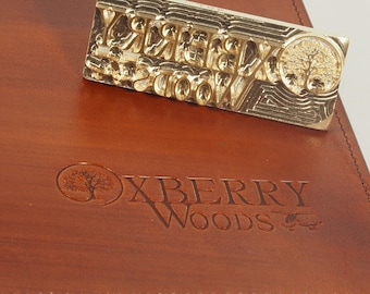 May include: A brown leather journal with a stitched edge and the embossed text "OXBERRY WOODS" and a tree logo. The journal is closed and lying on a white surface. A brass stamp with the same logo is lying on top of the journal.