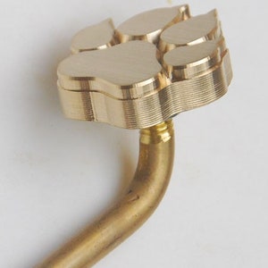 May include: A gold-colored metal pipe with a paw print-shaped end. The pipe is bent at a 90-degree angle.