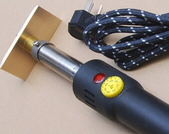 May include: A black electric heat press with a gold-colored metal plate attached to the end. The heat press has a temperature control knob and a red indicator light. It is plugged into a black and white cord.