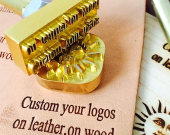 May include: A gold-colored metal stamp with the text "on leather on wood" and a heart-shaped design. The stamp is on a light brown surface with the text "Custom your logos on leather, on wood".