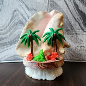 May include: A large, smooth, cream-colored conch shell serves as a backdrop for two miniature green plastic palm trees. The base features a marbled, multi-colored stone platform accented with small red and green coral-like pieces on a white stand.