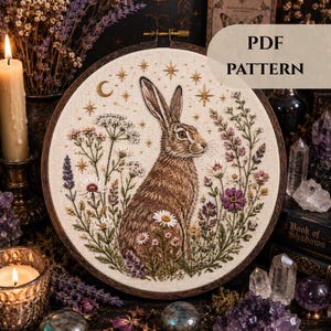 May include: An embroidered artwork in a wooden hoop, depicting a brown hare amidst wildflowers and stars. The design includes a crescent moon and the text "PDF PATTERN". The piece has a natural, rustic aesthetic, with detailed stitching and a woodland theme.