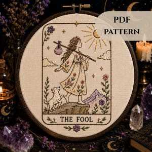 May include: An embroidery hoop containing a fabric panel with a hand-stitched design of The Fool tarot card. The image features a person walking with a staff and bundle, a small dog, a sun, and mountains. The text THE FOOL is stitched at the bottom.