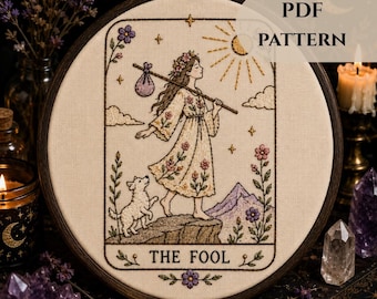 May include: An embroidery hoop containing a fabric panel with a hand-stitched design of The Fool tarot card. The image features a person walking with a staff and bundle, a small dog, a sun, and mountains. The text THE FOOL is stitched at the bottom.