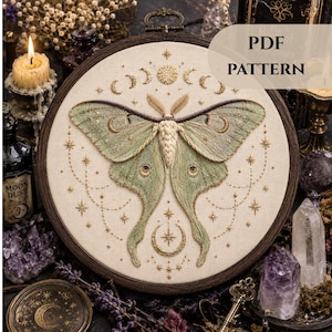 May include: A detailed embroidery hoop art piece featuring a large moth with green and gold wings, surrounded by celestial designs. The artwork is set against a cream-colored fabric, framed in a dark wooden hoop. The text "PDF PATTERN" is visible.