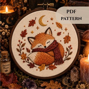 Peut inclure: Un tambour à broder contenant une pièce de tissu avec le motif brodé d'un renard endormi portant une écharpe rouge foncé, entouré de feuilles d'automne, de baies, d'étoiles et d'un croissant de lune. Le texte PDF PATTERN est visible en haut.