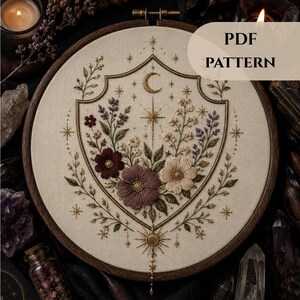 Può includere: Un telaio da ricamo contenente un tessuto color crema con un disegno a scudo ricamato raffigurante una mezzaluna, stelle e vari fiori nelle tonalità bordeaux, malva e crema. Il testo PDF PATTERN è visibile nell'angolo in alto a destra.