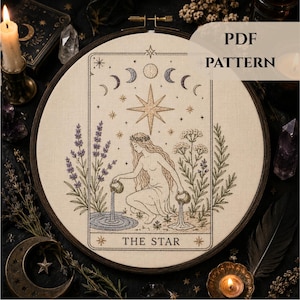 May include: An embroidery hoop containing a cream-colored fabric with a stitched design of The Star tarot card. The image features a figure pouring water from two pitchers, surrounded by lavender and yarrow plants, moon phases, and a large star.