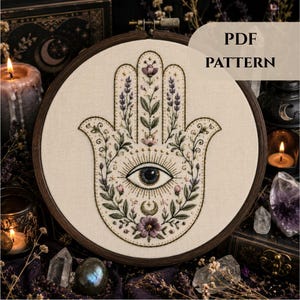 Könnte beinhalten: Ein Stickrahmen mit cremefarbenem Stoff und einem handgestickten Hamsa-Hand-Motiv. Das Design zeigt ein zentrales Auge, florale Elemente, Lavendelzweige und eine Mondsichel in den Farben Violett, Grün und Schwarz. Der Text lautet PDF PATTERN.