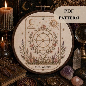 Op de afbeelding: Een kruissteek borduurring met een gedetailleerd ontwerp van de tarotkaart Rad van Fortuin. Het ontwerp bevat een centraal wiel, bloemelementen, een zon, maan en de tekst "THE WHEEL". De woorden "PDF PATTERN" zijn ook zichtbaar.