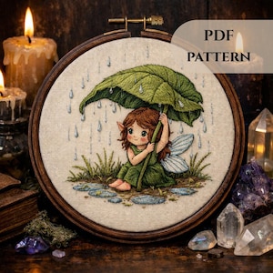 Peut inclure: Broderie encadrée représentant une fée sous un parapluie en forme de feuille, sous la pluie. La fée a les cheveux bruns et porte une robe verte. Le texte "PDF PATTERN" est visible sur l'ouvrage.