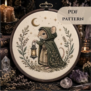 Op de afbeelding: Een borduurring met een stoffen paneel met een geborduurde illustratie van een muis in een donkergroene cape met capuchon die een lantaarn vasthoudt. Het ontwerp bevat een maansikkel, sterren en bloemenkransen. Tekst leest PDF PATTERN.
