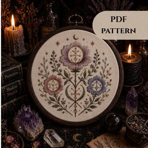 Könnte beinhalten: Ein runder Stickrahmen mit einem floralen Design aus lila, rosa und blauen Blumen, einem Mond und Sternen. Der Text "PDF PATTERN" ist zu sehen. Der Rahmen befindet sich vor einem dunklen, mystischen Hintergrund mit Kerzen und Kristallen.