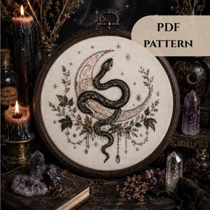 May include: A circular embroidery hoop containing a fabric panel with a stitched design of a dark snake coiled around a crescent moon, surrounded by botanical elements and small stars. The text PDF PATTERN is visible in the upper right corner.