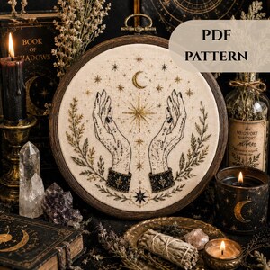 Peut inclure: Un tambour à broder présentant un tissu crème avec des broderies en fil noir et doré représentant deux mains tendues vers un croissant de lune et des étoiles, entourées de tiges botaniques. Le texte PDF PATTERN est visible en haut à droite.