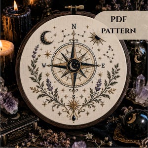 May include: An embroidery hoop containing a cream-coloured fabric with a compass rose design in navy blue and gold thread. The pattern features a central crescent moon, cardinal directions, stars, and floral sprigs. The text PDF PATTERN is visible.