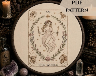 May include: An embroidery hoop containing a fabric panel with a stitched tarot card design titled THE WORLD. The design features a central figure surrounded by a floral wreath, with a human head, eagle, bull, and lion in the corners. Text reads PDF PATTERN.