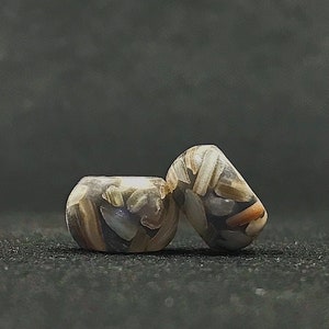 May include: Two barrel-shaped beads featuring a mosaic-like pattern of embedded fragments in shades of tan, brown, gray, and white. The beads have a smooth, polished finish with a central hole running through the middle of each cylindrical form.