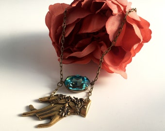 Puede incluir: Un collar de estilo vintage con un colgante de mano en tono bronce y un gran cristal ovalado de color turquesa. El collar está suspendido de una delicada cadena. Una rosa de color coral en el fondo.