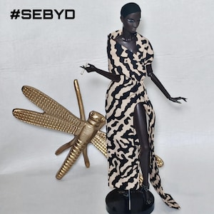 May include: A dark-skinned fashion doll wearing a beige and black abstract patterned wrap dress with a high leg slit, standing next to a large gold-colored dragonfly figurine. The text #SEBYD is visible in the upper left corner of the image.