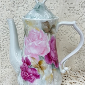 May include: White porcelain teapot with a lid, featuring hand-painted pink and magenta roses with green and yellow leaves. The teapot has a curved spout and handle, and a decorative finial on the lid.