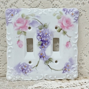 May include: A white ceramic double light switch plate featuring a hand-painted floral design with pink roses and purple lilac clusters. The rectangular plate has a decorative embossed border and four screw holes for mounting on a wall.