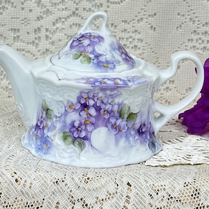 May include: A white porcelain teapot featuring a hand-painted floral design of purple violets with green leaves. The teapot has a scalloped base, a curved handle, a pointed spout, and a matching lid with a small handle on top.