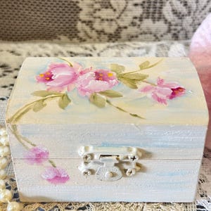 May include: A small, rectangular wooden box with a hinged lid. The box is painted light blue with pink floral accents and a white metal clasp. The flowers are pink with yellow centers and green leaves. The box is likely used for jewelry or small keepsakes.