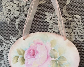 Puede incluir: Adorno de pared ovalado pintado a mano con una rosa rosa y hojas verdes. La obra de arte está sobre un fondo rosa claro con un lavado azul pálido. Se adjunta una cinta rosa para colgar.
