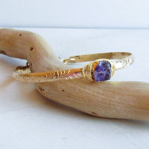 May include: A gold bangle bracelet with a purple gemstone set in the center. The bracelet is on a piece of driftwood.