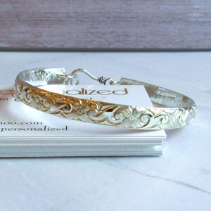 May include: Silver bangle bracelet with a detailed floral pattern. The open bracelet has a hook clasp. The bracelet is displayed on a stack of white cards, with the word "alized" visible.