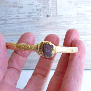 May include: Gold bangle bracelet with a dark brown, rough-cut gemstone set in the center.