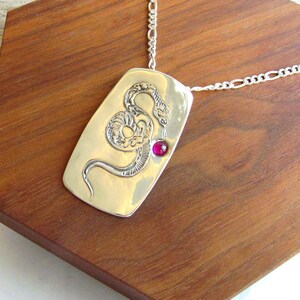 May include: A silver necklace with a rectangular pendant featuring a detailed snake design and a small red gemstone. The necklace is displayed on a wooden stand.