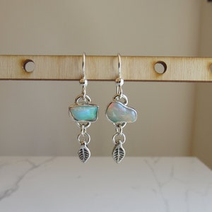 May include: A pair of silver earrings with a teardrop-shaped opal stone and a leaf charm hanging from each earring.