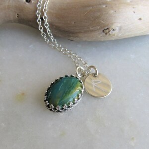 May include: A silver chain necklace with a green and gold oval pendant set in a silver crown design. The pendant is attached to a silver charm with the letter 'R' engraved on it.