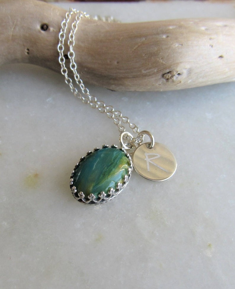 May include: A silver chain necklace with a green and gold oval pendant set in a silver crown design. The pendant is attached to a silver charm with the letter 'R' engraved on it.