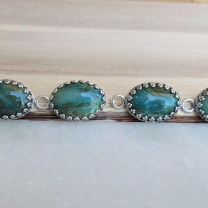 May include: A silver chain necklace with six oval-shaped turquoise and green gemstone pendants. Each pendant is set in a silver bezel with a decorative border.