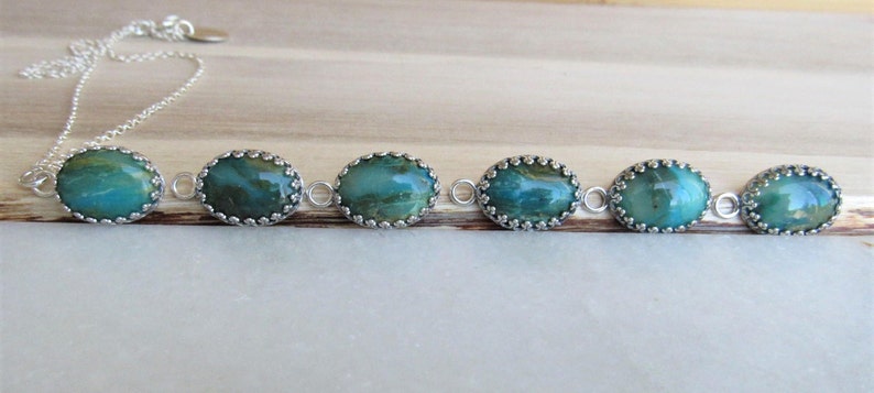 May include: A silver chain necklace with six oval-shaped turquoise and green gemstone pendants. Each pendant is set in a silver bezel with a decorative border.