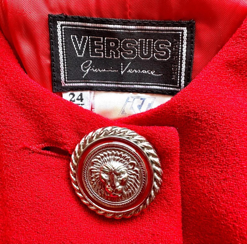 Puede incluir: Primer plano de una prenda roja con un bot&oacute;n adornado con la cabeza de un le&oacute;n. La etiqueta dice "VERSUS Gianni Versace" e incluye una etiqueta de talla 24. La tela tiene un aspecto texturizado.