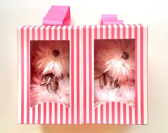 May include: Two rectangular boxes with pink and white vertical stripes, each featuring a clear window displaying a fluffy light pink plush dog toy. Each toy wears a dark brown ribbon around its neck with the text Juicy Couture printed on it.
