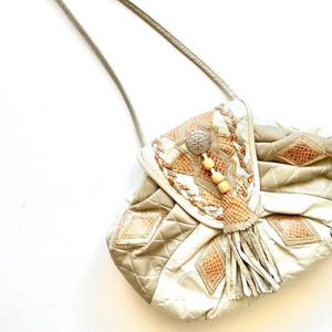 May include: A vintage beige and white quilted crossbody bag with a decorative flap. The flap features a braided design, a silver-tone button, wooden beads, and a tassel. The bag has diamond-shaped faux snakeskin accents.