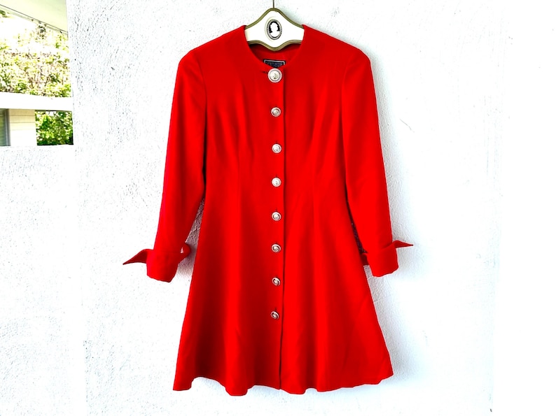 Puede incluir: Un abrigo-vestido rojo brillante con mangas largas y falda acampanada. El abrigo presenta una fila de botones grandes, redondos y plateados en la parte delantera y botones a juego en los pu&ntilde;os. El abrigo est&aacute; colgado en una percha.
