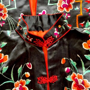 Puede incluir: Una bata de seda negra con ribetes rojos y bordados florales. La bata tiene un cuello mandar&iacute;n y un cierre frontal con nudos decorativos.