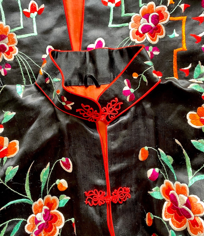 Puede incluir: Una bata de seda negra con ribetes rojos y bordados florales. La bata tiene un cuello mandar&iacute;n y un cierre frontal con nudos decorativos.